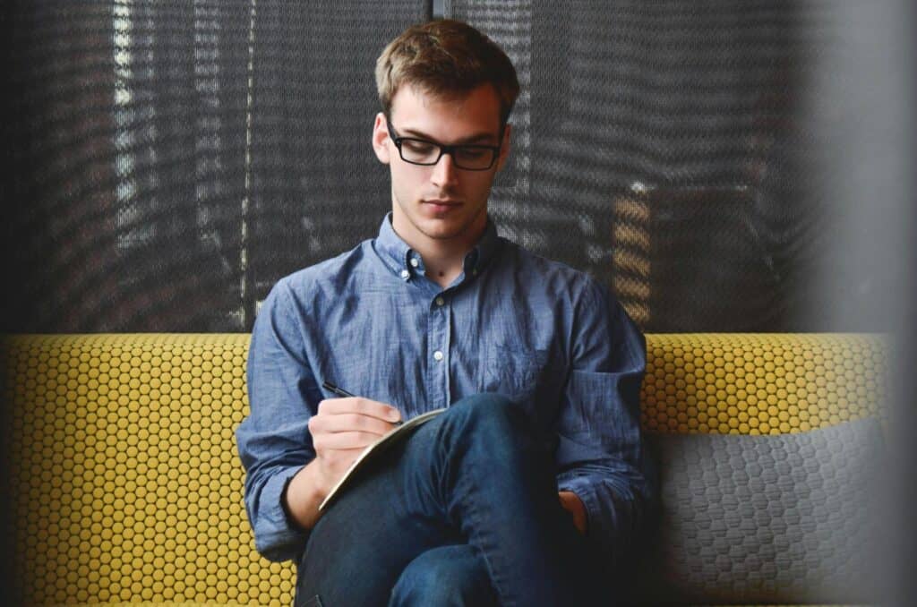 A young man sitting on a modern couch, focused on writing notes, representing active learning and self-study techniques for language improvement.